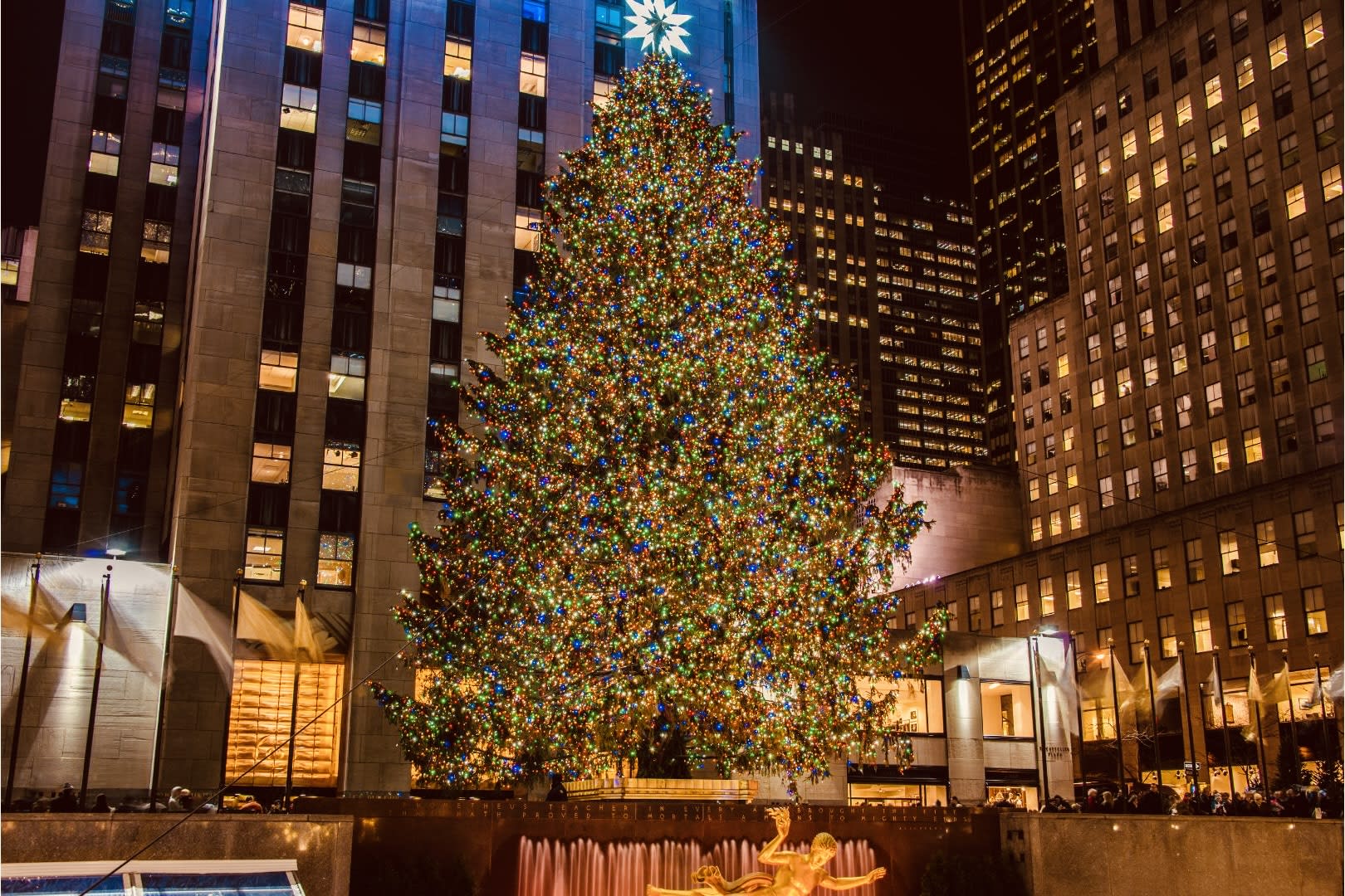 A large Christmas tree adorned with colorful lights stands proudly in a vibrant city plaza at night, representing one of the most iconic gay Christmas destinations. Tall buildings surround the festive scene, exuding a welcoming and inclusive atmosphere perfect for LGBTQ+ travelers seeking holiday joy. A golden statue is visible near the tree, reflecting in the dazzling lights and adding to the lively and festive spirit of this quintessential holiday adventure. This magical destination invites everyone to celebrate love, diversity, and unity during this special time of year.