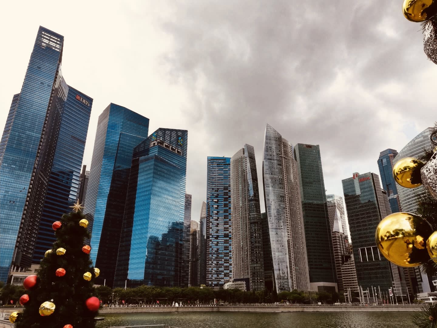 A cityscape featuring numerous modern skyscrapers with glass facades beneath a cloudy sky beautifully captures the festive spirit of one of the renowned gay Christmas destinations. In the foreground, part of a decorated Christmas tree adorned with red and gold ornaments is visible, showcasing this vibrant LGBTQ+ holiday hotspot.