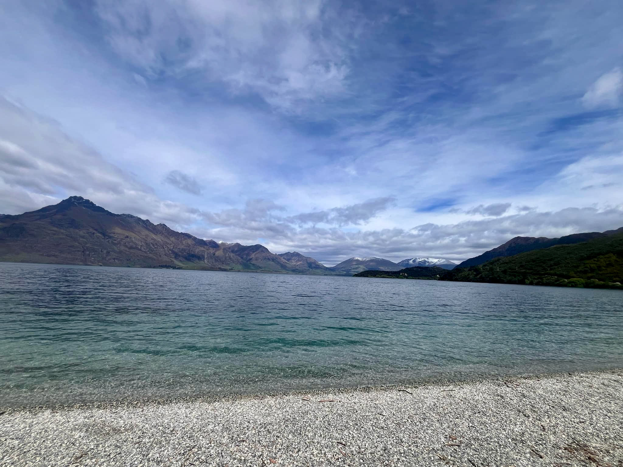 A serene lake with clear, calm water stretches across the foreground, meeting a rocky shoreline. In the distance, a range of snow-capped mountains under a partially cloudy sky creates a dramatic backdrop. This peaceful, natural scene evokes the beauty and allure of easy moves to safe havens abroad. Among these havens is New Zealand, one of the LGBTQ-friendly countries to move to as an American, offering not only breathtaking landscapes but also welcoming communities that embrace diversity and inclusivity.