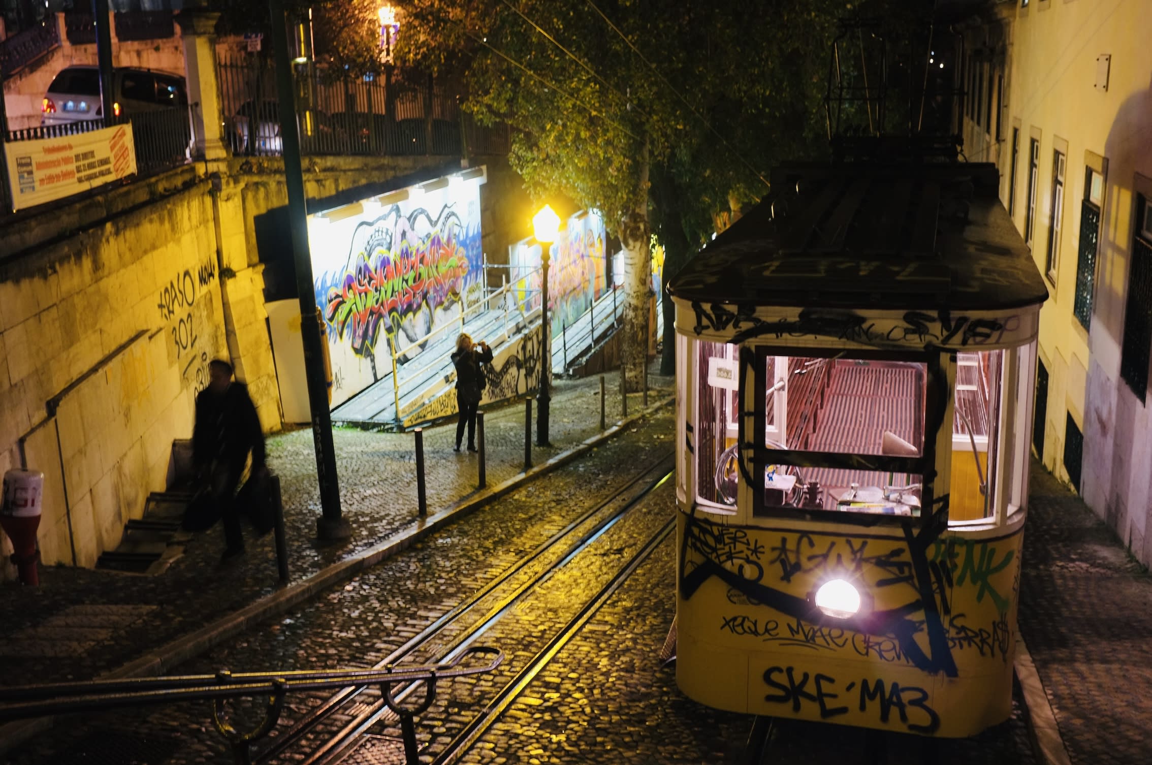 A nighttime scene unfolds as a tram ascends a graffiti-covered hill in one of the LGBTQ+-friendly countries to move to as an American. The cobblestone path is illuminated by streetlights, casting a warm glow on the surroundings with trees gently swaying overhead. Two people stand near the tracks, their silhouettes outlined against the urban landscape. A brightly lit mural bursts into vibrant color amidst the dim setting, hinting at this safe haven abroad—a testament to diversity and inclusion that these welcoming destinations promise for those seeking acceptance and community far from home.