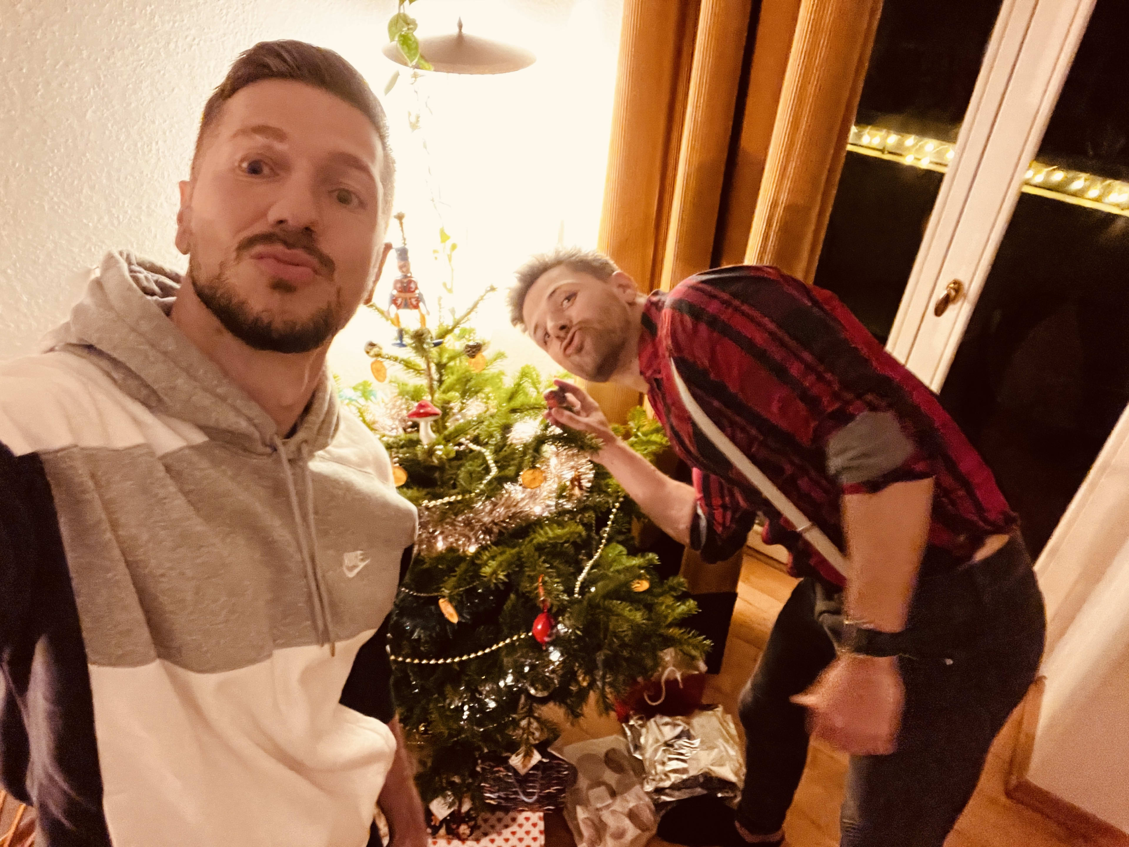 Two people are decorating a Christmas tree with lights and ornaments in a cozy room, capturing the festive spirit. As one snaps a selfie while the other adjusts decorations, they discuss their dream of exploring top gay Christmas destinations. Wrapped gifts lie under the tree, and warm lighting enhances the holiday cheer in this inviting setting, perfectly reflecting their excitement for unique holiday travels.