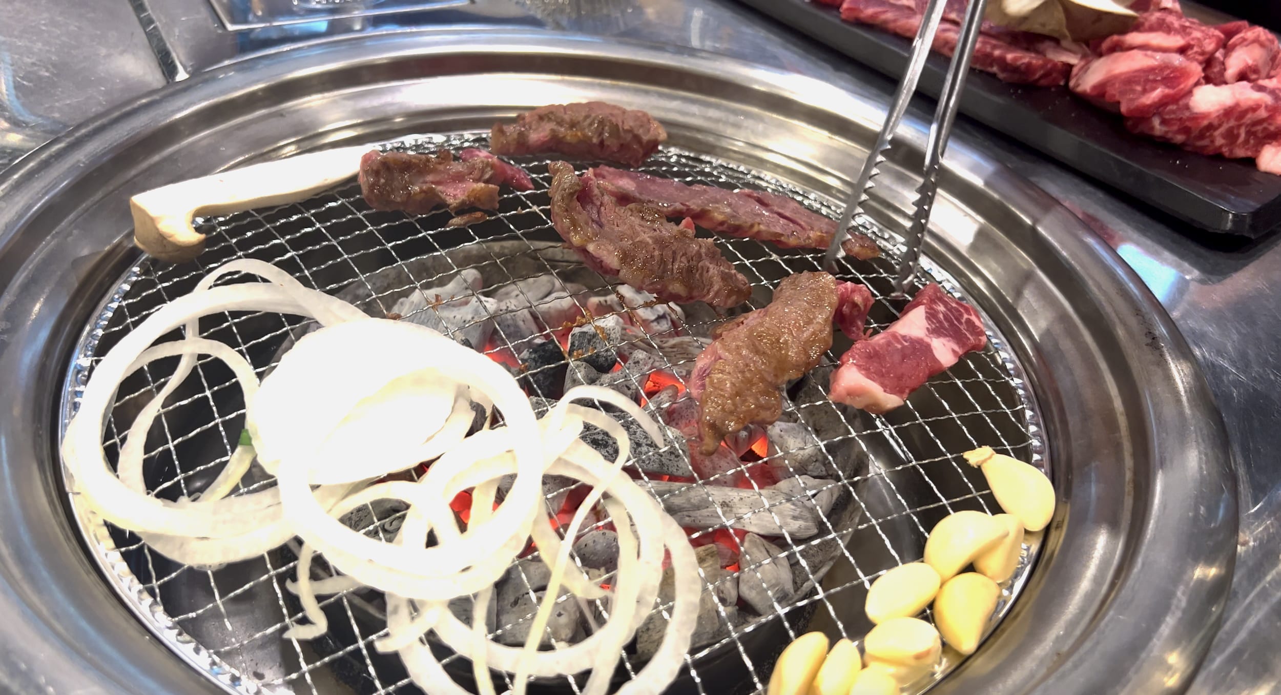 Slices of meat, onions, and garlic sizzle on a grill over hot coals, offering a taste of authentic cuisine reminiscent of the vibrant streets you might explore in one day in Seoul.