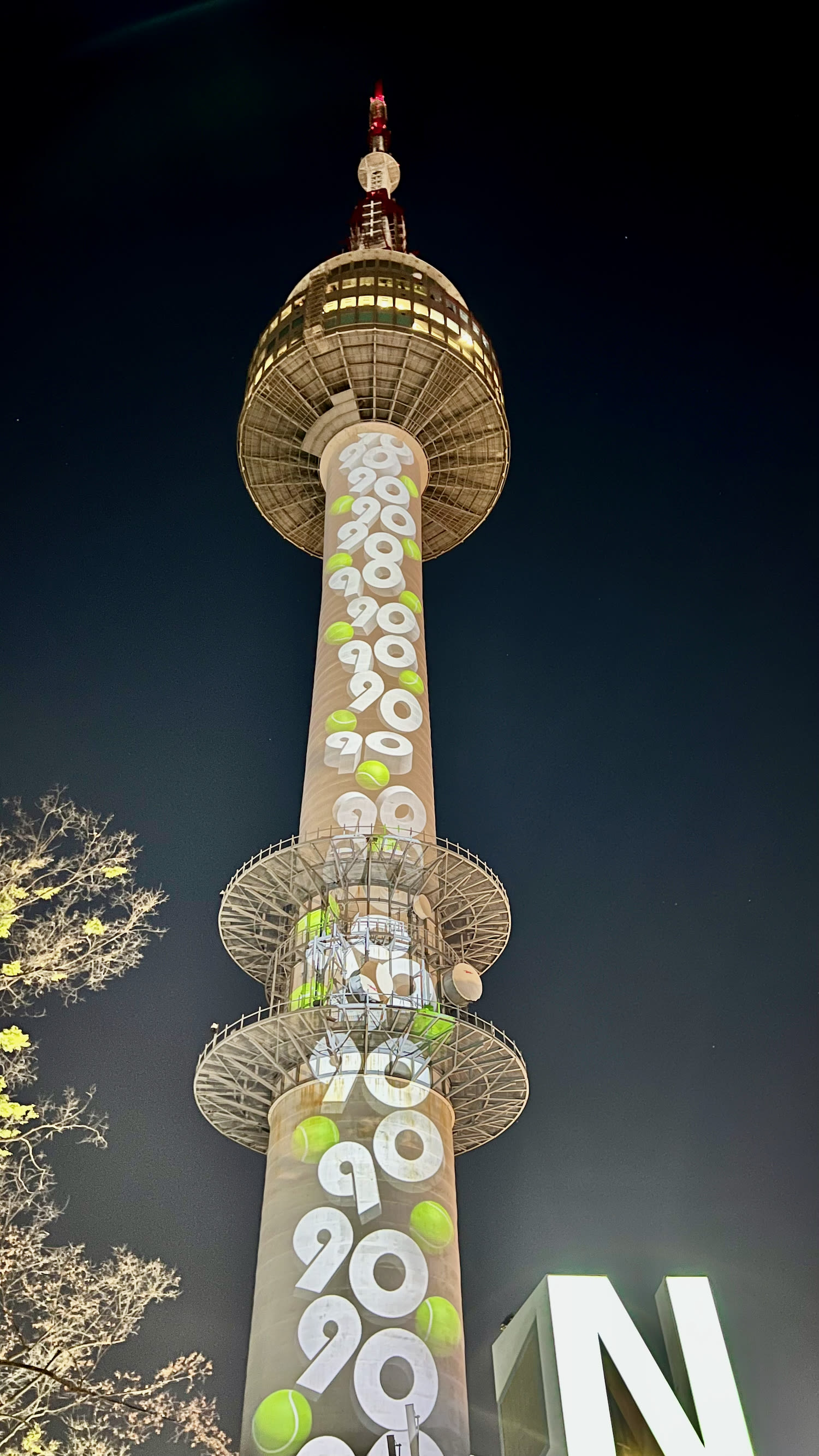 A tall tower with circular patterns stands illuminated against the night sky in Seoul, featuring numbers and tennis ball designs. If you have just one day in Seoul, this stunning structure is a must-see, rivaling the city's iconic landmarks.