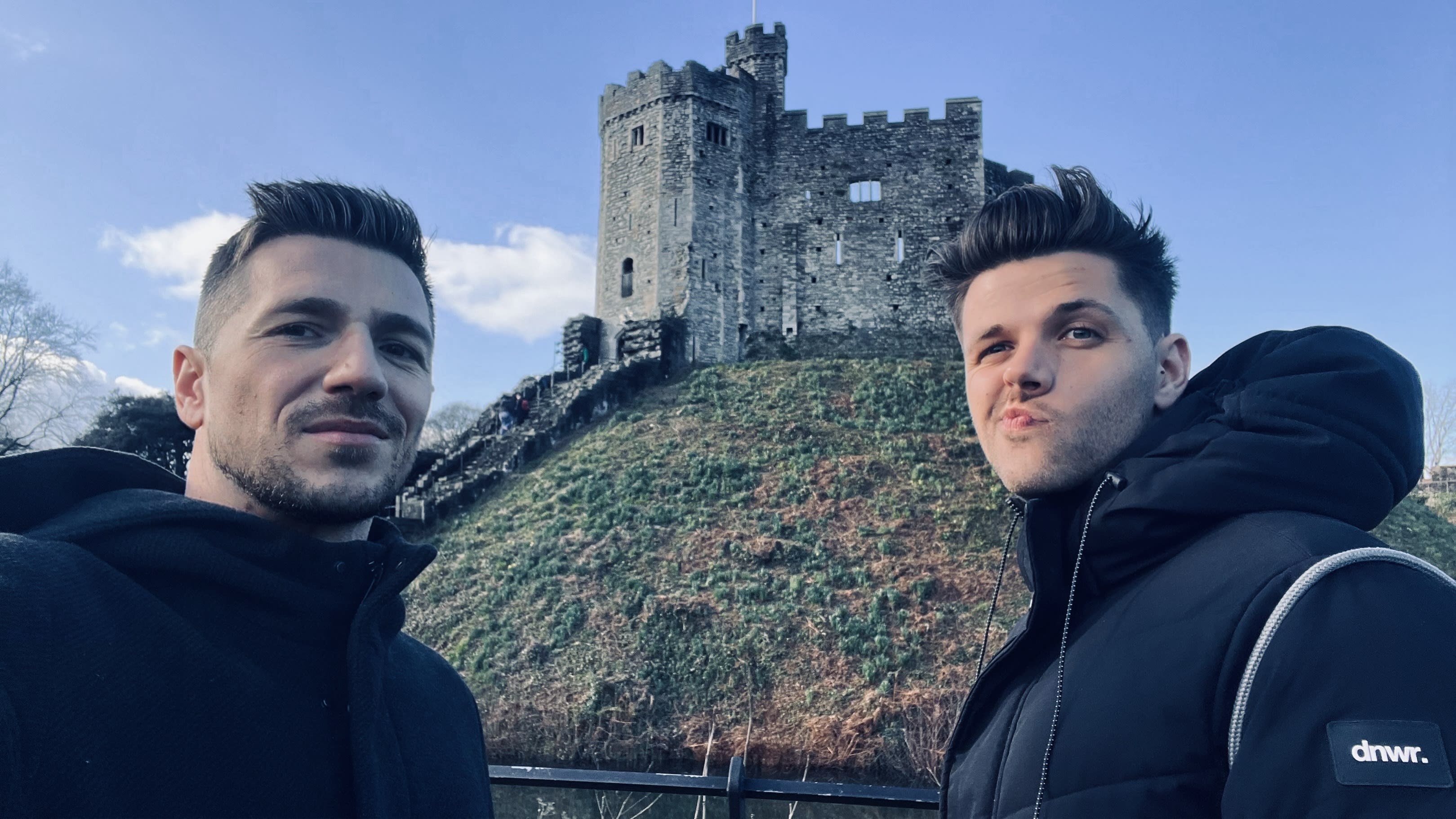 Two people are posing in front of a stone castle on a grassy hill, set against the clear sky of Wales. This moment captures just one highlight of their adventurous Wales itinerary, which promises breathtaking landscapes and rich history at every step.