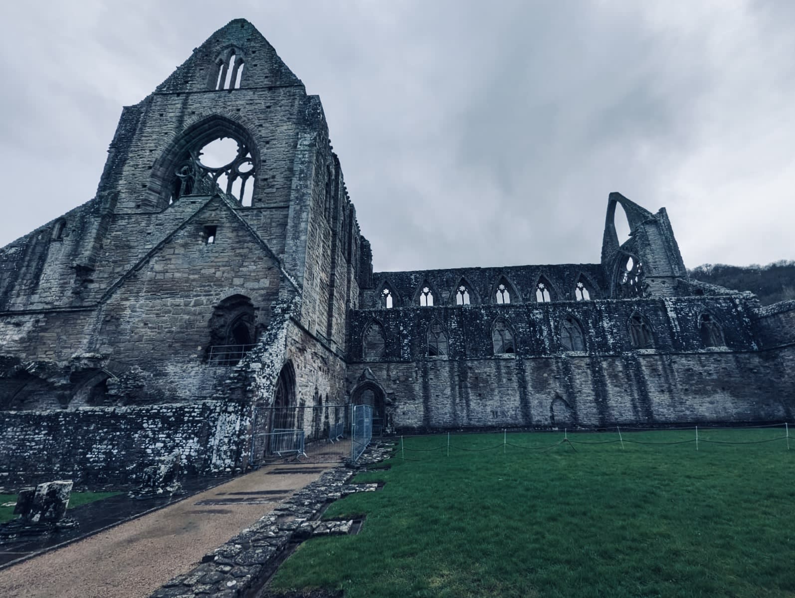 A large, weathered stone abbey ruins stand under a cloudy sky in Wales, with an arched window and grassy foreground. This captivating scene invites you to explore a mesmerizing wales itinerary, whispering tales of hidden cottages and distant sandy shores.