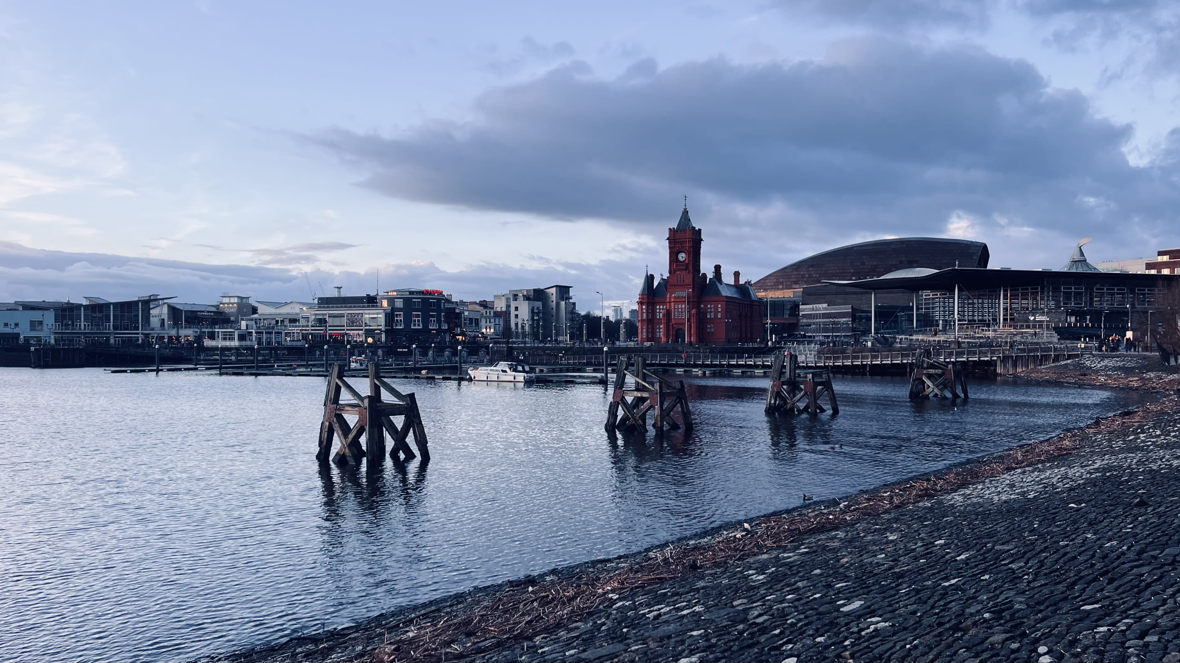 A waterside view of a cityscape in Wales, ideal for a Wales itinerary, features a red brick building and wooden posts in the water, contrasted by a modern structure with a curved metal roof under the cloudy sky. These stunning views are reminiscent of hidden cottages and offer visitors unique sights to explore.