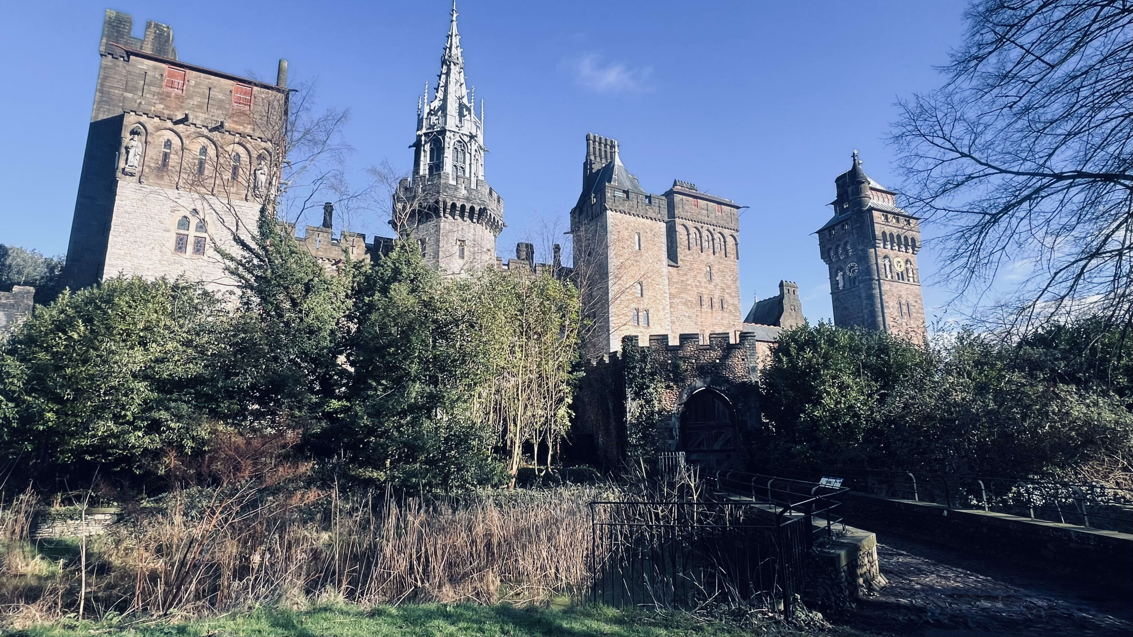 Nestled in the heart of Wales, the historic castle with stone towers and spires stands majestically against a clear blue sky, surrounded by hidden cottages and lush trees. Don't miss this breathtaking sight on your Wales itinerary, as it perfectly captures the enchanting essence of Welsh heritage and natural beauty.