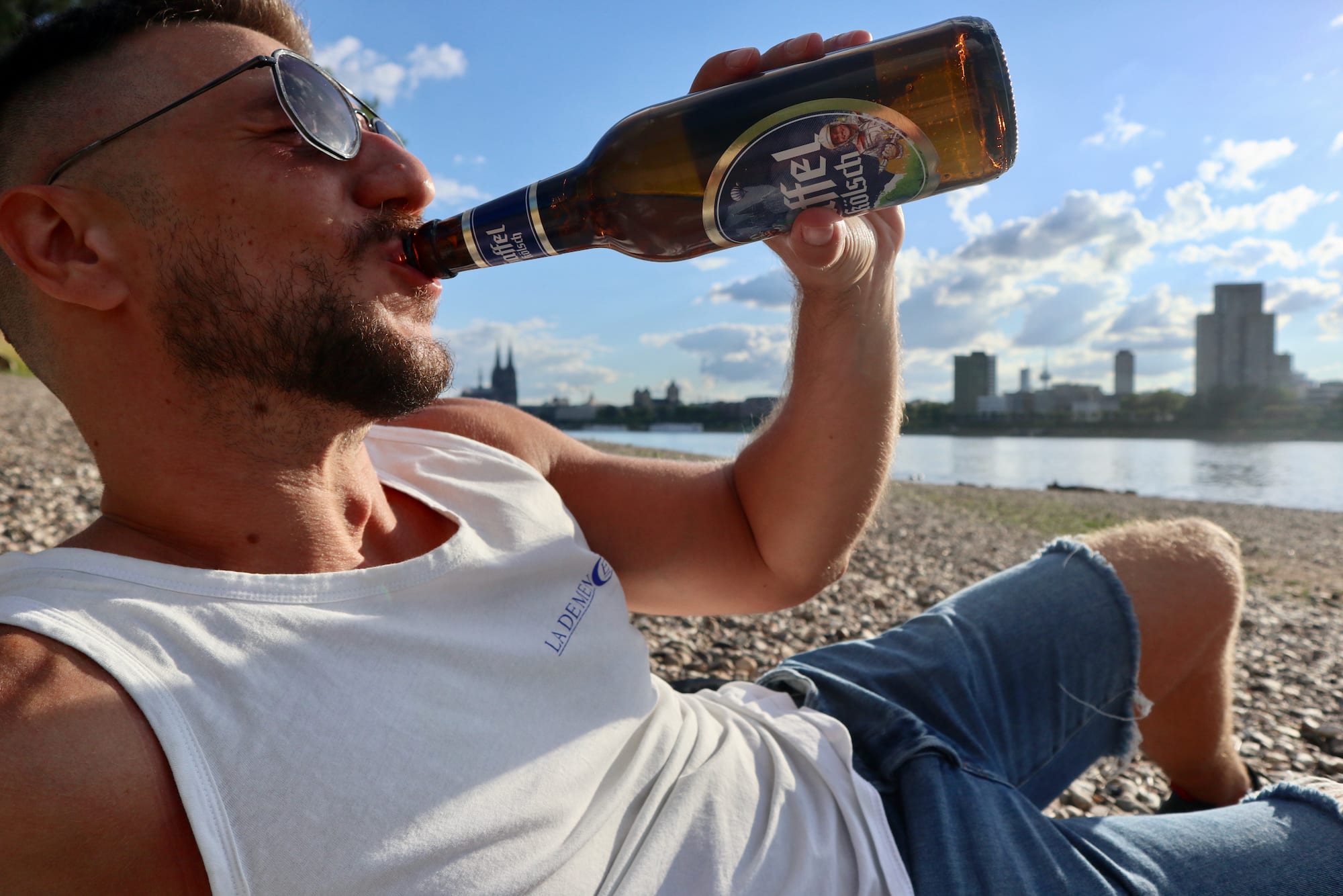 A man wearing sunglasses and a tank top sips from a beer bottle while lounging on a pebble beach beside the river, soaking in the lively atmosphere of gay Cologne. The vibrant city skyline serves as an intriguing backdrop. This moment captures the essence of hot gay romance novels, where picturesque settings enhance irresistible connections.