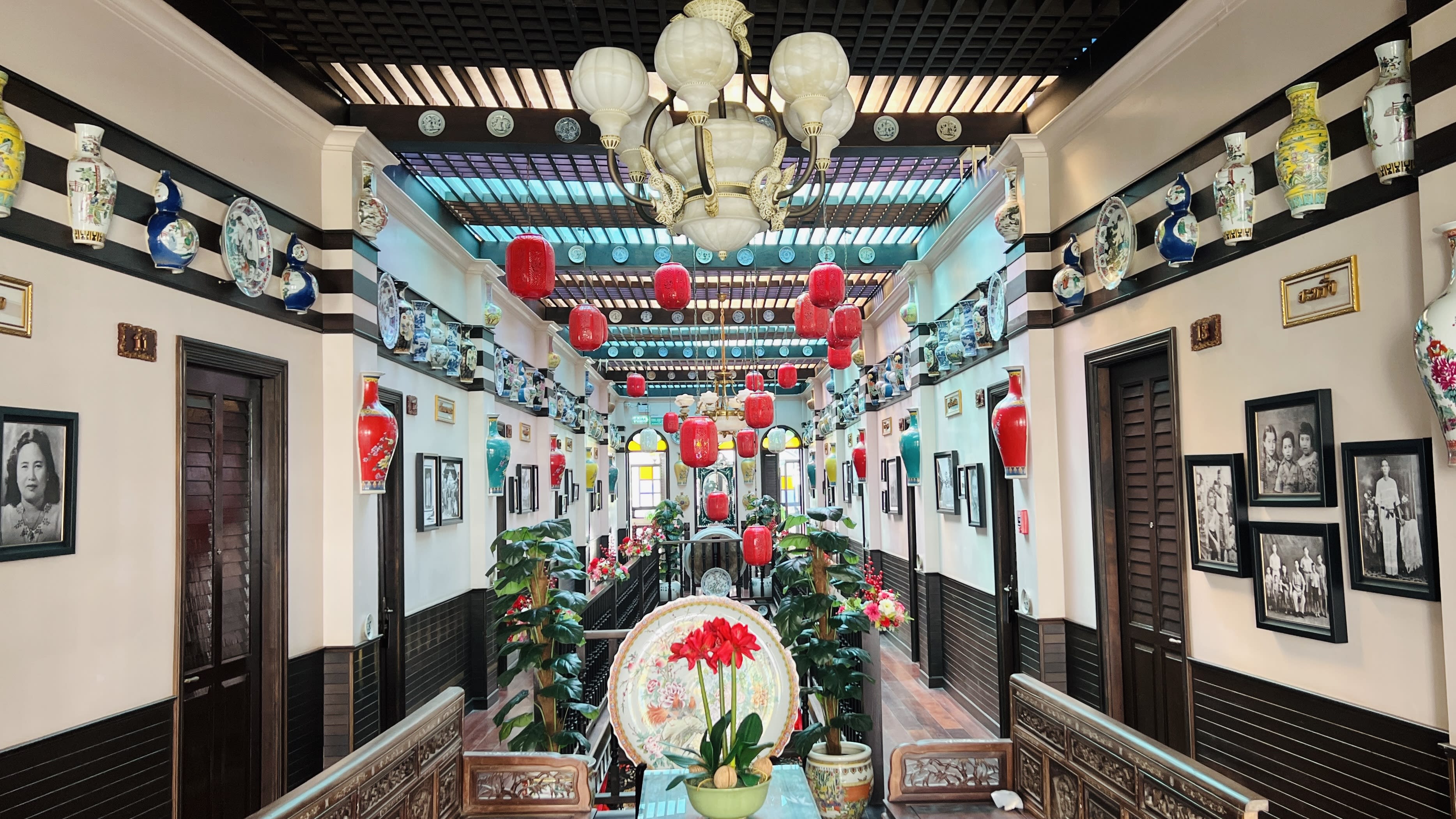 This traditional interior exudes historic charm with red lanterns and framed photos adding a nostalgic touch. Decorative vases and lush plants enrich the setting, while a chandelier illuminates the scene. A large plate with a flower design prominently displayed captures the cultural allure of Songkhla Old Town, where such captivating elements offer a glimpse into its rich heritage.