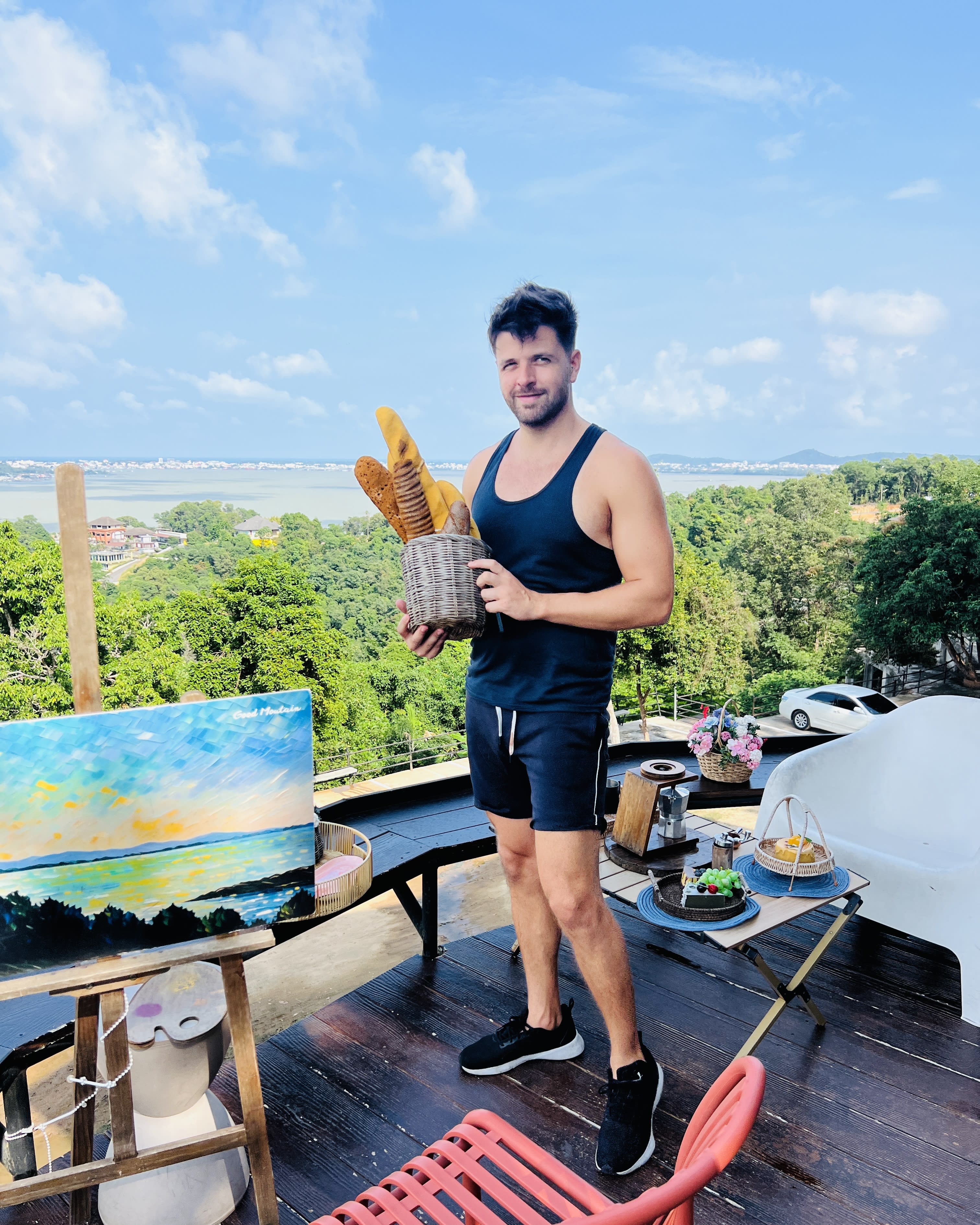 A man in a black tank top and shorts stands on a deck, holding a basket of baguettes. Nearby, captivating street art enhances the outdoor seating area. The scenic view, framed by trees and sky, evokes the historic charm of Songkhla Old Town. This harmonious blend of culture and nature offers an enchanting glimpse into the area's unique character and beauty.