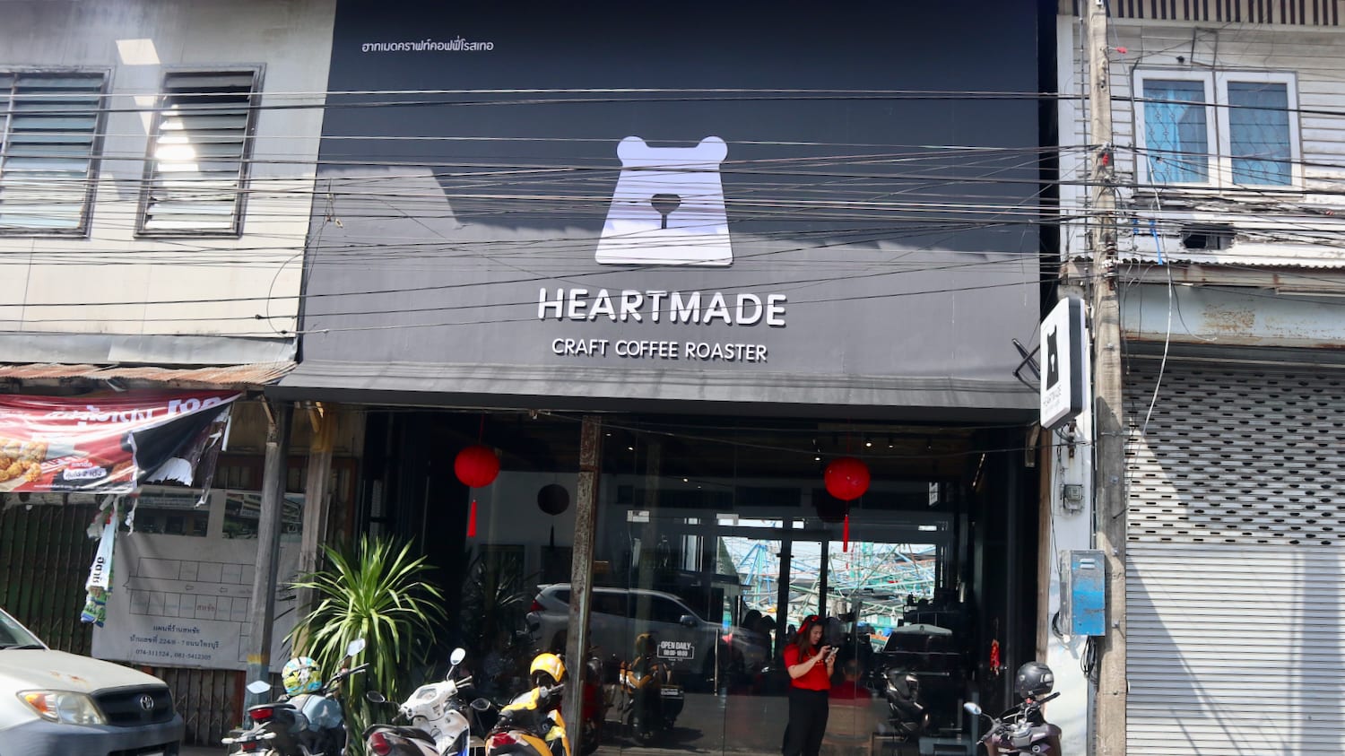Street view of Heartmade Craft Coffee Roaster in Hat Yai, with its charming bear logo prominently displayed. Motorcycles line the front, suggesting its popularity among locals and travelers. A person can be seen entering this queer-friendly cafe, known for welcoming the LGBTQ+ community. It's a vibrant spot for those exploring gay Hat Yai and seeking great coffee and atmosphere.