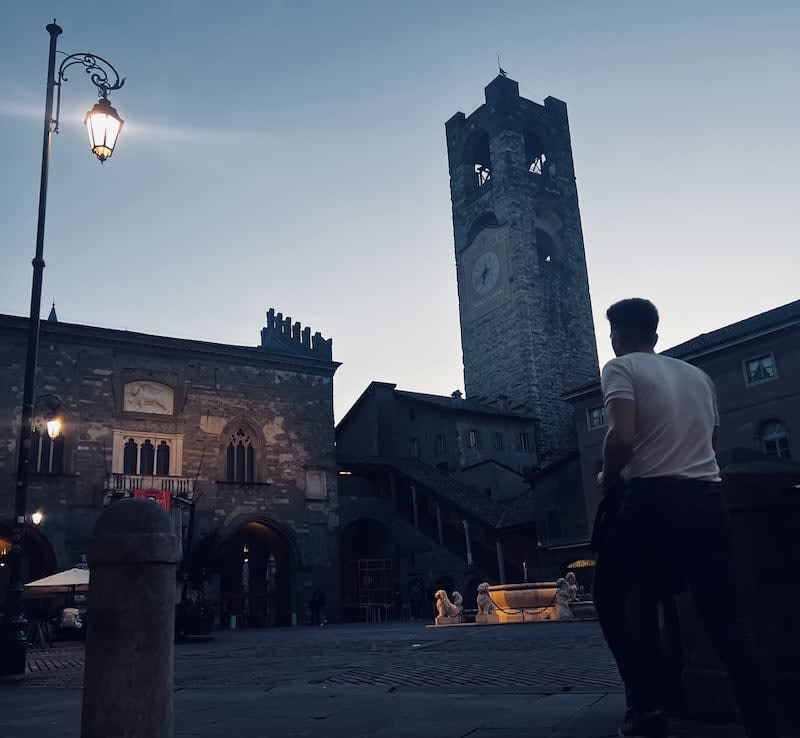 A man stands in a plaza at dusk, gazing at a historic stone building with a tall clock tower. Lit street lamps and cozy seating areas evoke the charm of Venetian history, ideal for those savoring authentic bites on their 2 day Venice itinerary. The scene captures the essence of Venice, offering travelers memorable moments to enhance their journey through this enchanting city.
