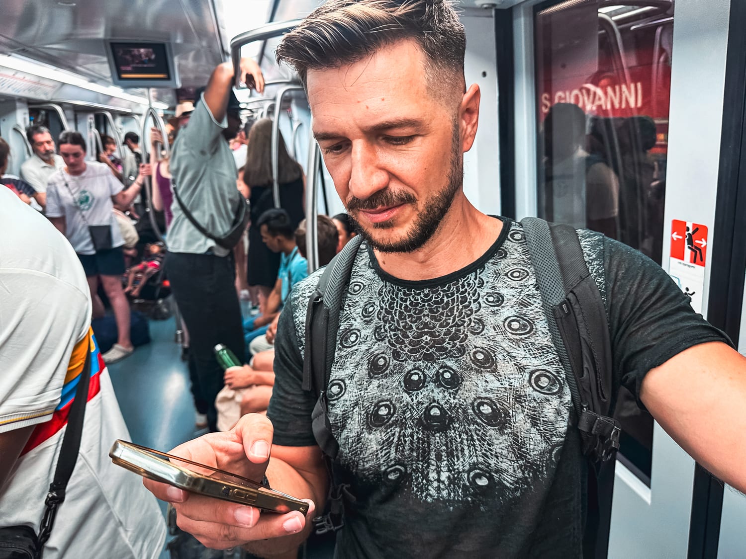 A man with a backpack stands in a crowded subway train, looking at his smartphone, likely checking mobile data. Other passengers are seated and standing, creating a busy commuting atmosphere.