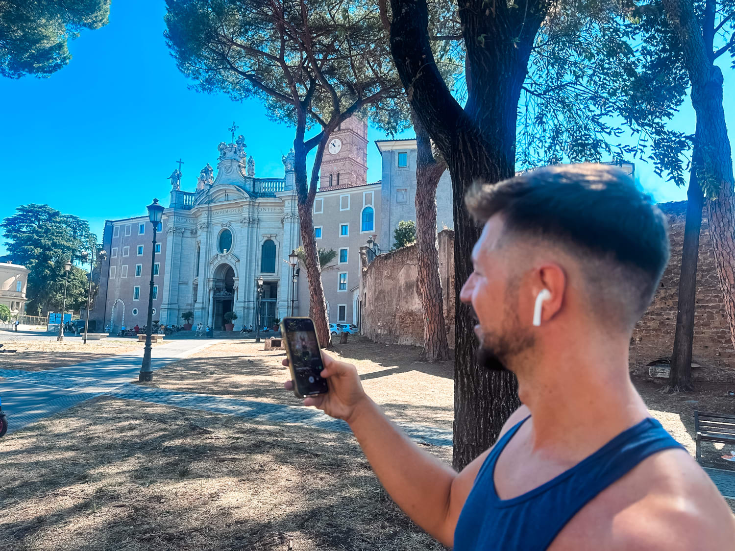 A man in a tank top and earbuds snaps a photo by a historic building, enjoying sunny Europe trips with the best esims for europe.