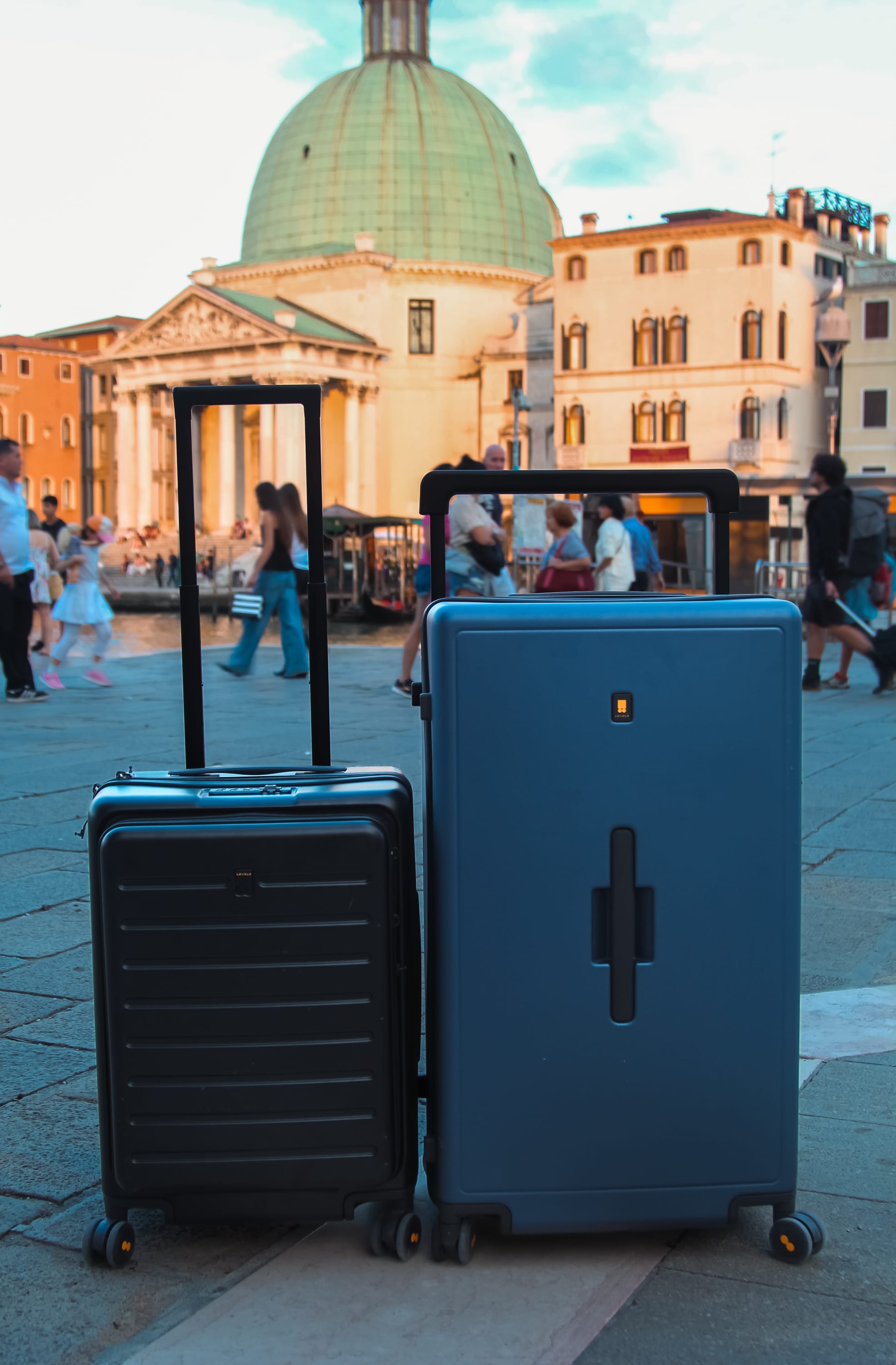 Two durable suitcases, one black and one blue, stand upright on a stone plaza. People walk by in the background, and a domed historic building is visible in the distance—an ideal scene for digital nomads exploring a European city.