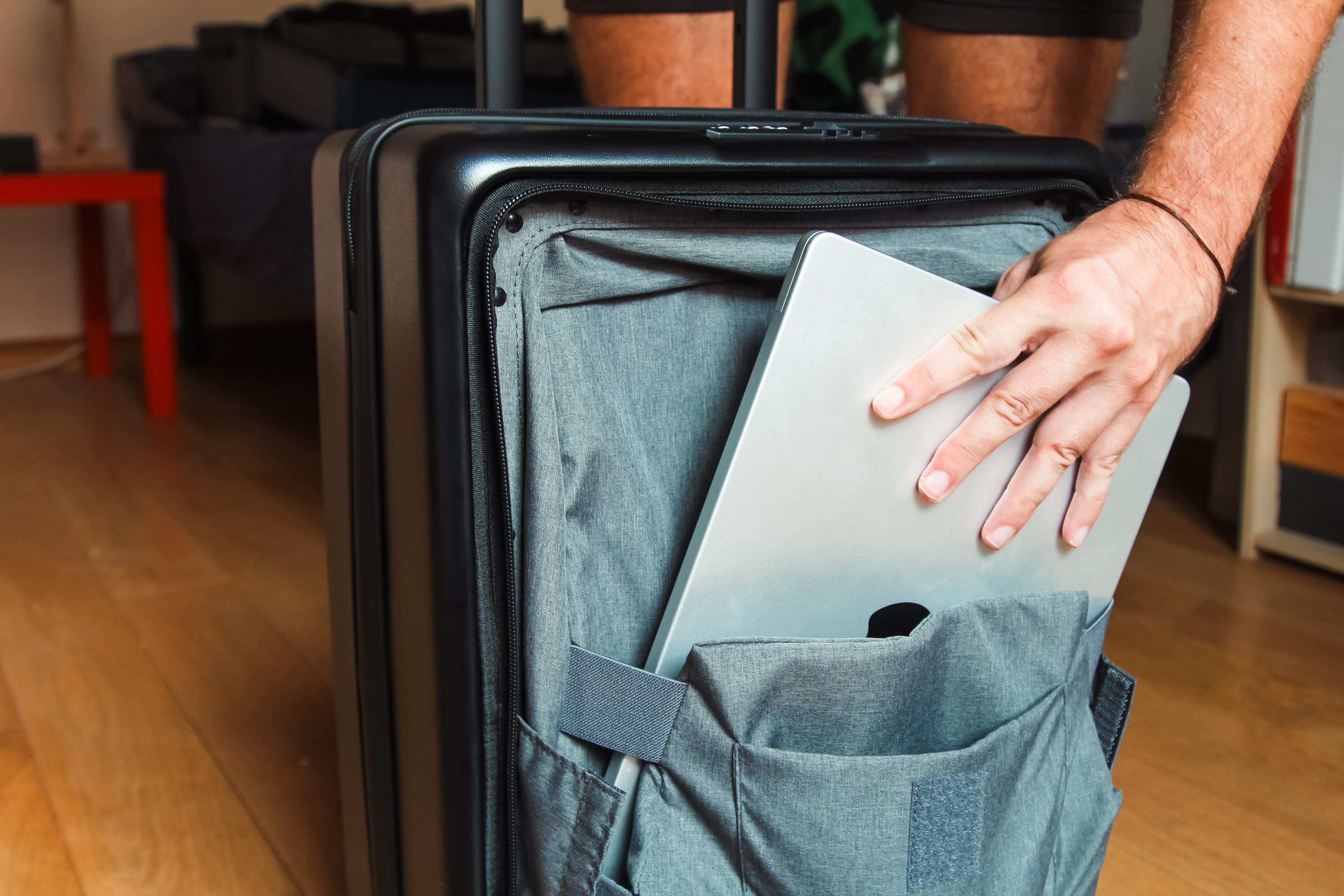 A traveler places a closed laptop into the front pocket of a LEVEL8 Road Runner, one of the most durable suitcases, which stands upright on a wooden floor with a red table visible in the background.