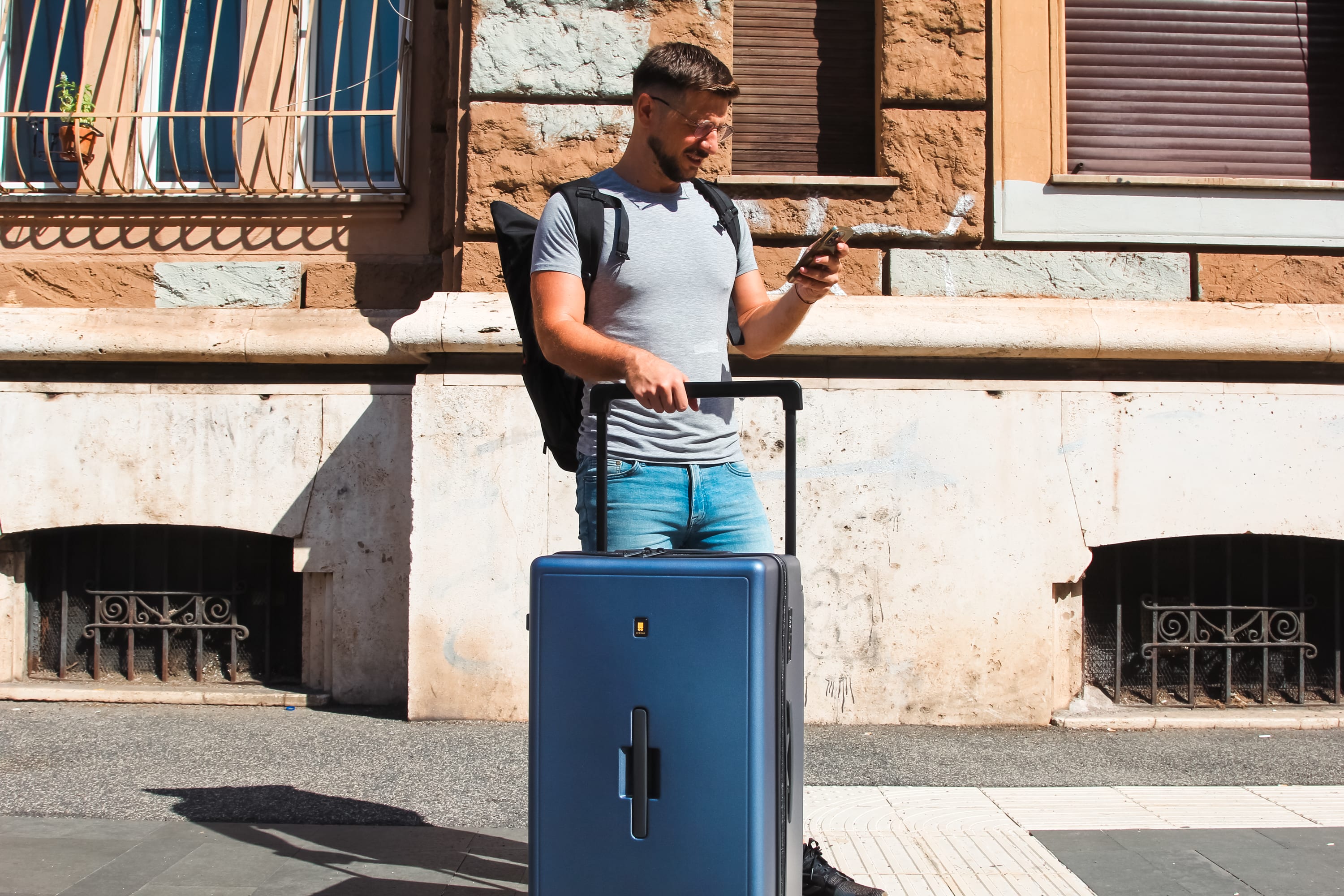 A man stands on a sidewalk in front of a building, holding the handle of a LEVEL8 Voyageur suitcase and checking his phone. Dressed casually with a backpack, he captures the spirit of digital nomads who rely on durable suitcases for their journeys.