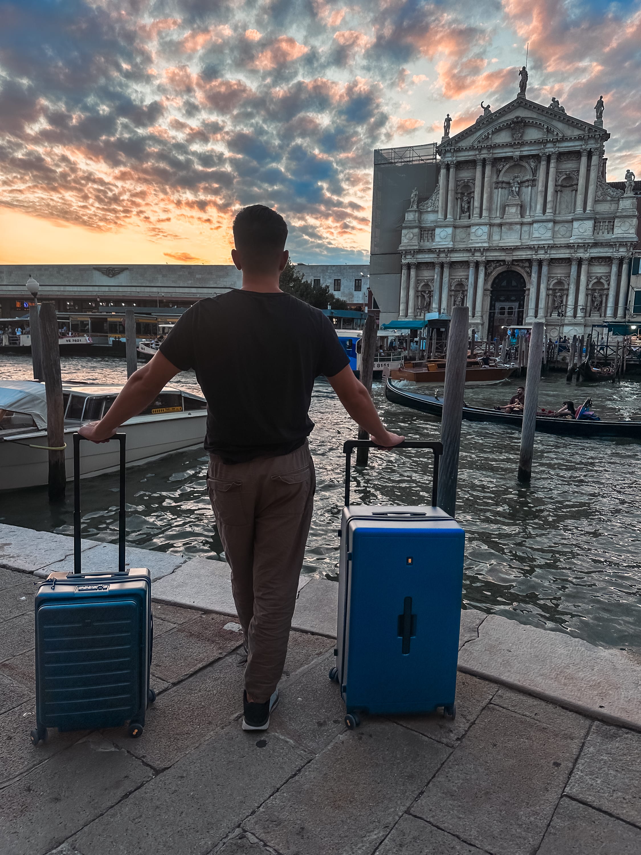 A person stands on a stone walkway by a canal, holding LEVEL8 suitcases, with boats drifting in the water and a historic building glowing in the sunset—perfect travel luggage for digital nomads exploring new destinations.