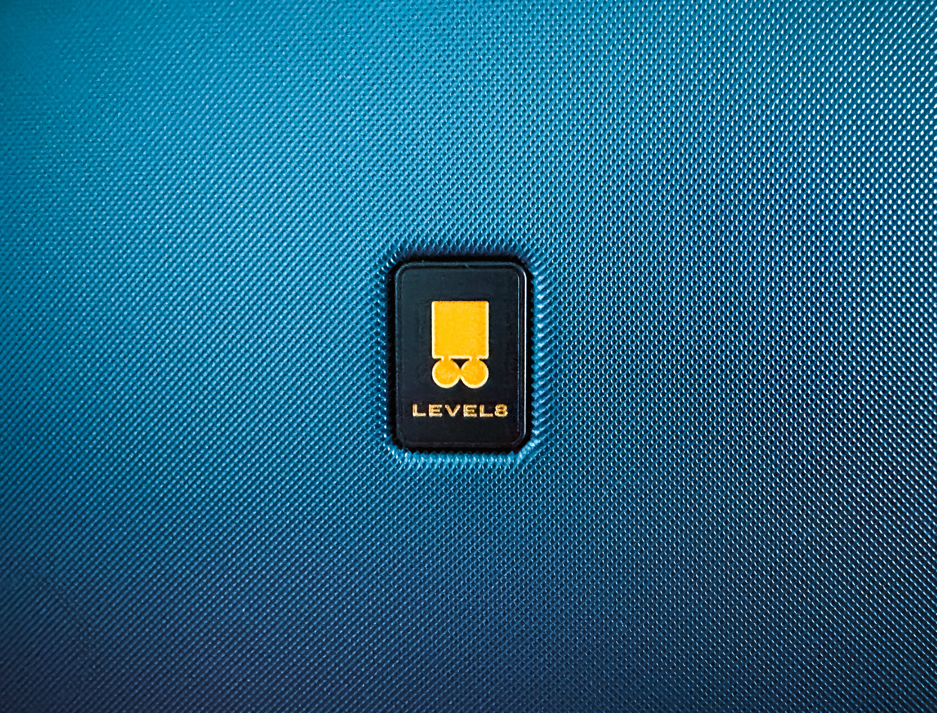 Close-up of a LEVEL8 Voyageur luggage logo on a blue textured surface. Perfect for travelers, the durable suitcase features a yellow square and circle above 'LEVEL8' in white, displayed on a black rectangular patch.