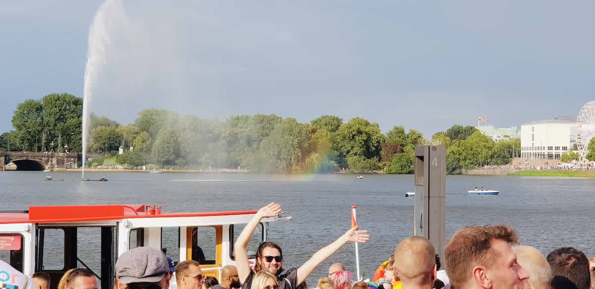 A spectacular view of a rainbow created by a water fountain during a sunny gay pride day in Hamburg, with joyful attendees in the foreground, symbolizing hope and inclusion.