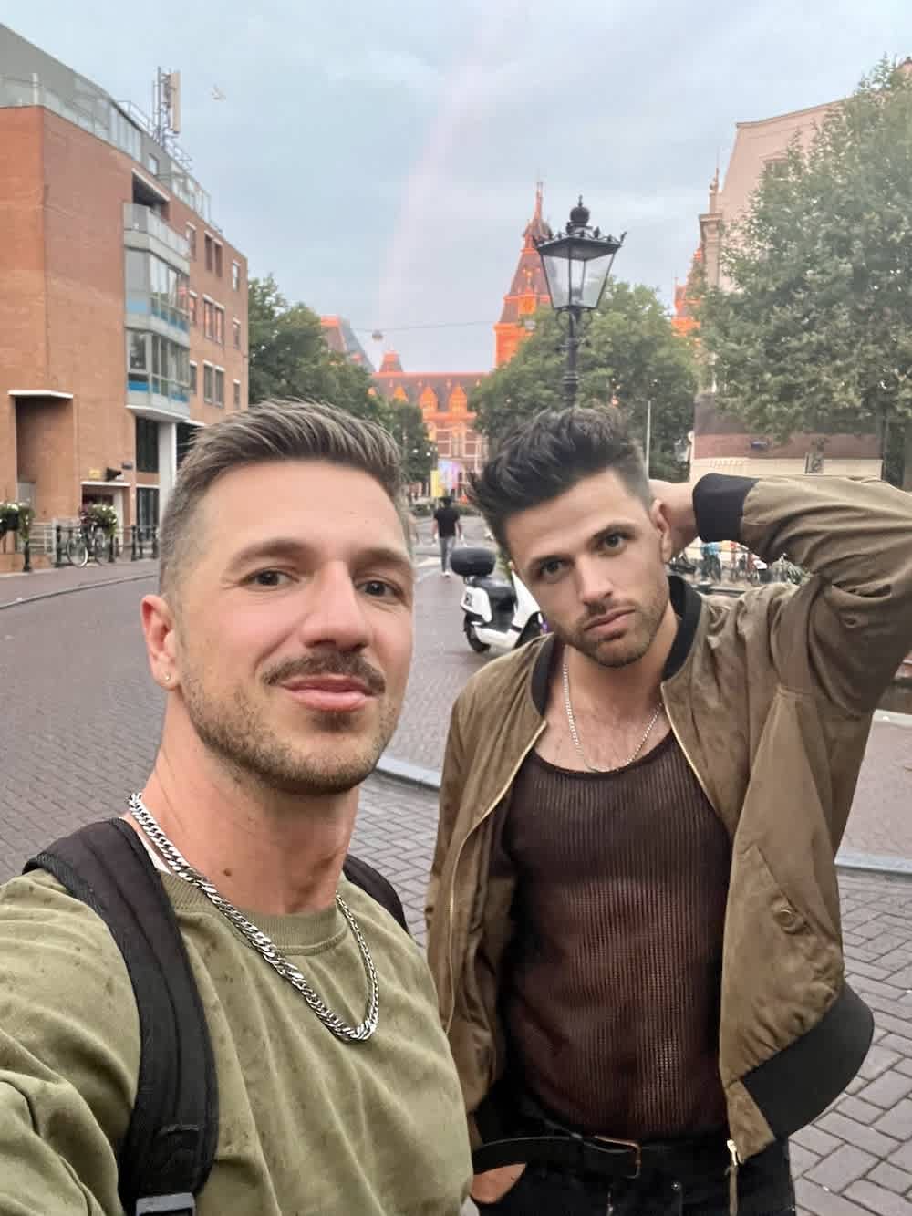 Two men stand confidently in front of a historical European Amsterdam street, illustrating the personal freedom and unity celebrated during gay pride events across Europe.