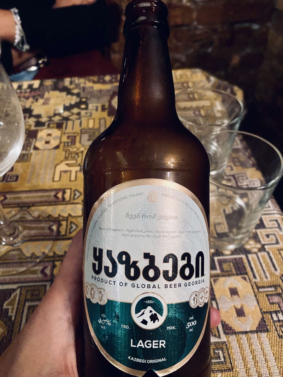 A hand holding a bottle of Kazbegi Original Lager, a product of Global Beer Georgia, showcasing the authentic Georgian beer against a backdrop of traditional patterned fabric.