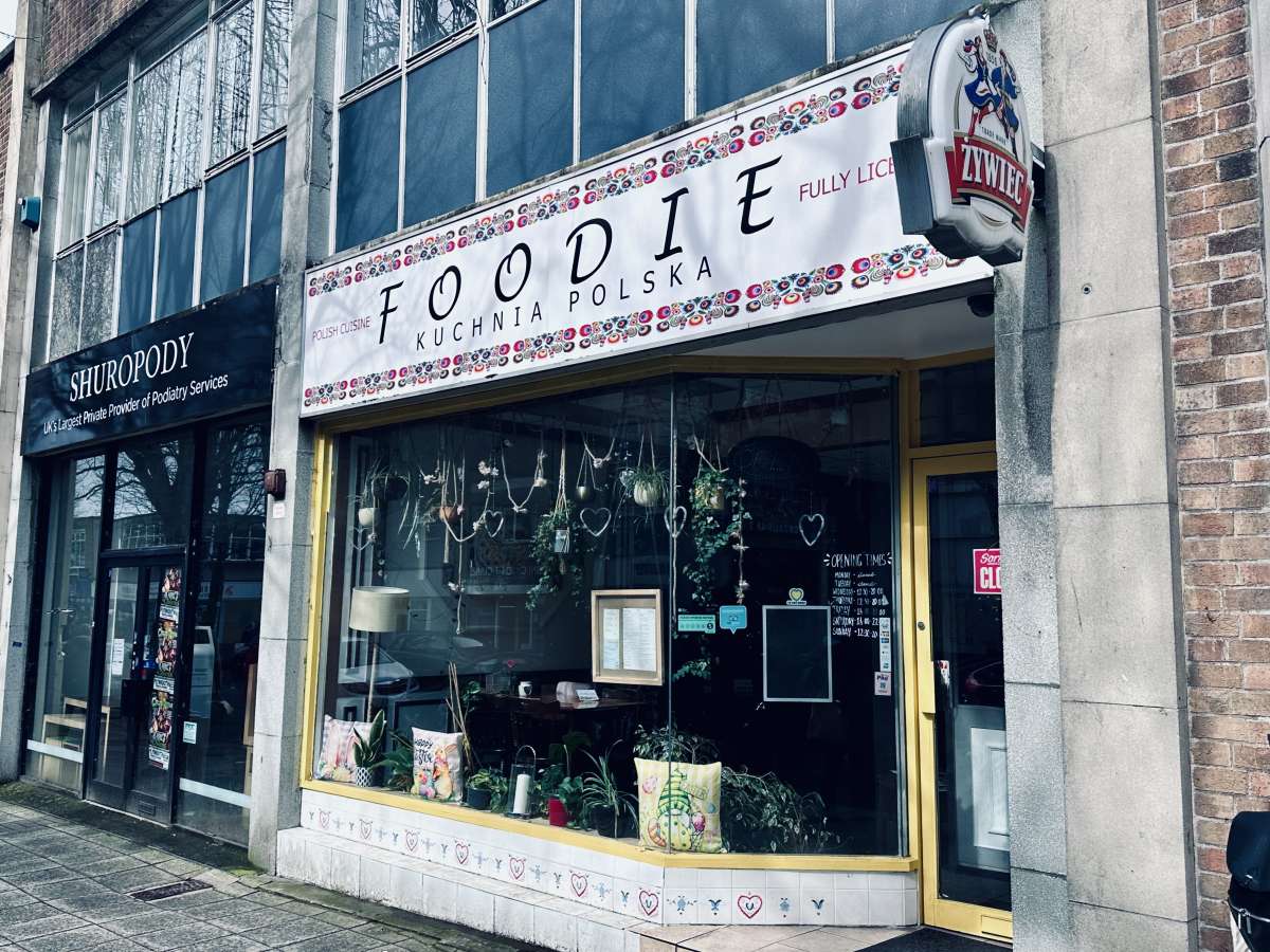 Exterior view of FOODIE Kuchnia Polska, a Polish cuisine restaurant in Plymouth, with colorful folk patterns decorating its signboard, next to Shuropody foot clinic, symbolizing the diverse commercial offerings of the city.