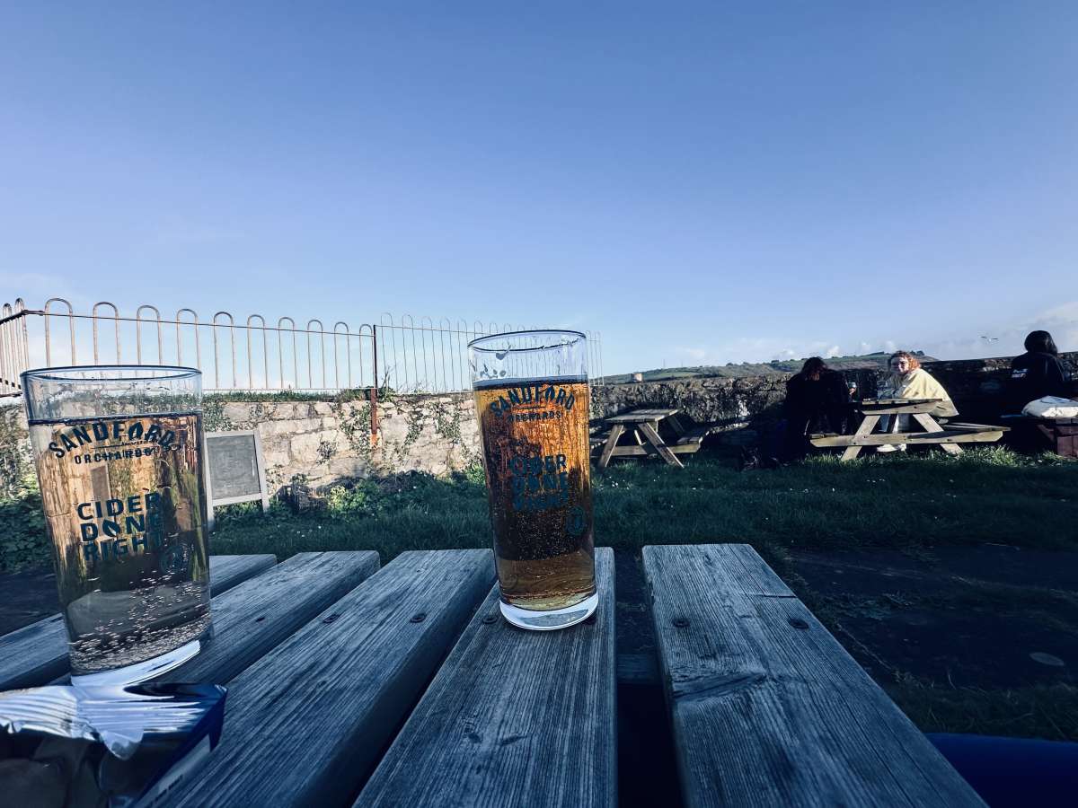 A refreshing perspective from Dutton's Cafe in Plymouth, featuring two glasses of Sandford Orchards cider on a wooden picnic table with patrons enjoying the outdoor seating area under a clear blue sky.