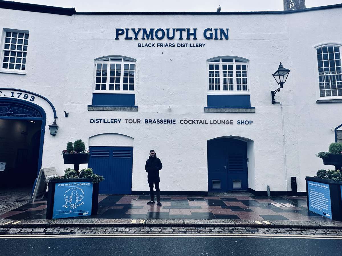 Front view of the historic Plymouth Gin Black Friars Distillery building, with its bold signage and deep blue doors, as a visitor stands in front, and informational signs about The Barbican welcome passersby.