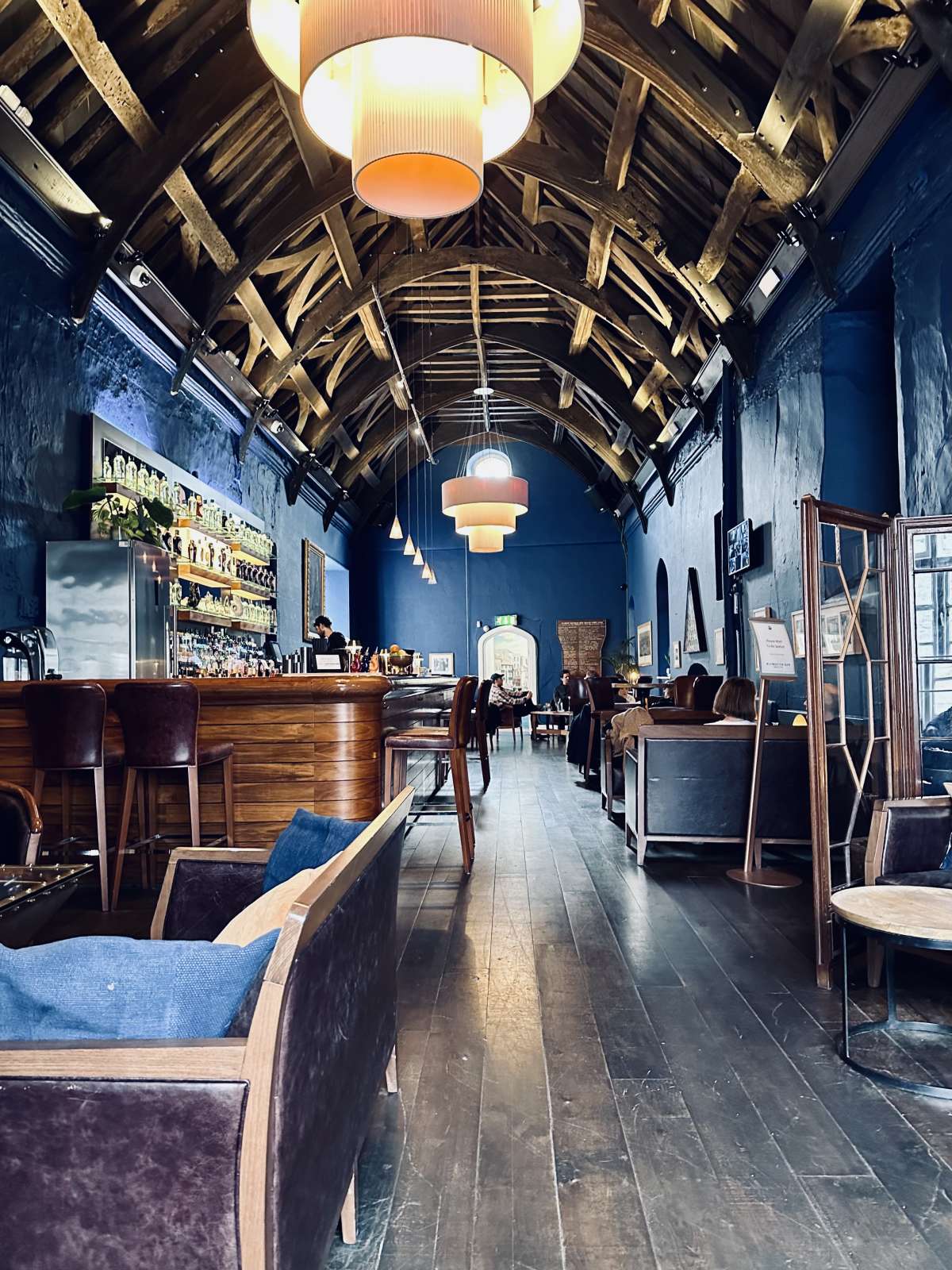 Interior of the Plymouth Gin Distillery bar with a rustic aesthetic, featuring high wooden beam ceilings, cozy blue upholstered seating, and warm pendant lighting, inviting a relaxed ambiance.