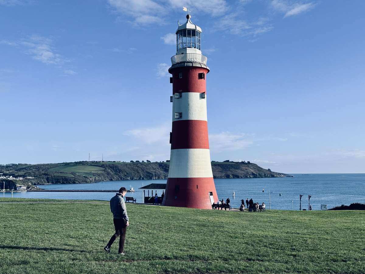 A clear day at Plymouth Hoe, showcasing the iconic Smeaton's Tower lighthouse with its red and white stripes, as a person strolls by and others enjoy the scenic ocean view in the background.