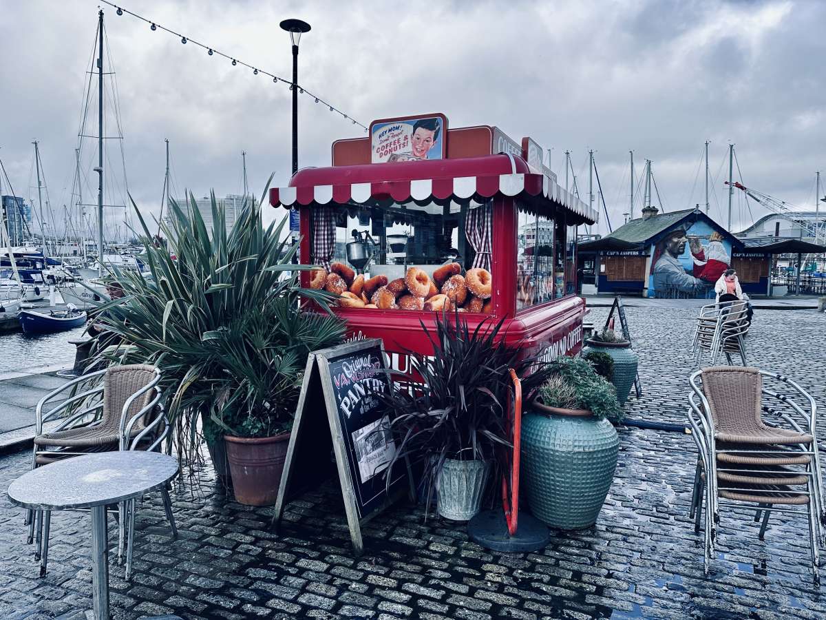 A cozy doughnut stand with a red and white striped awning offers a sweet treat at Plymouth's marina, with boats moored in the background under a grey sky, capturing a relaxed waterfront vibe.