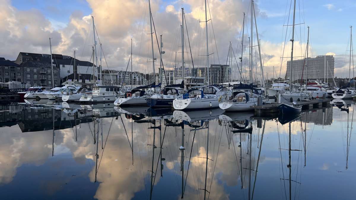 Calm waters at the Plymouth marina reflect an array of moored sailboats and yachts at dusk, with the urban skyline gently silhouetted against a cloud-streaked sky, encapsulating a serene harborside evening.