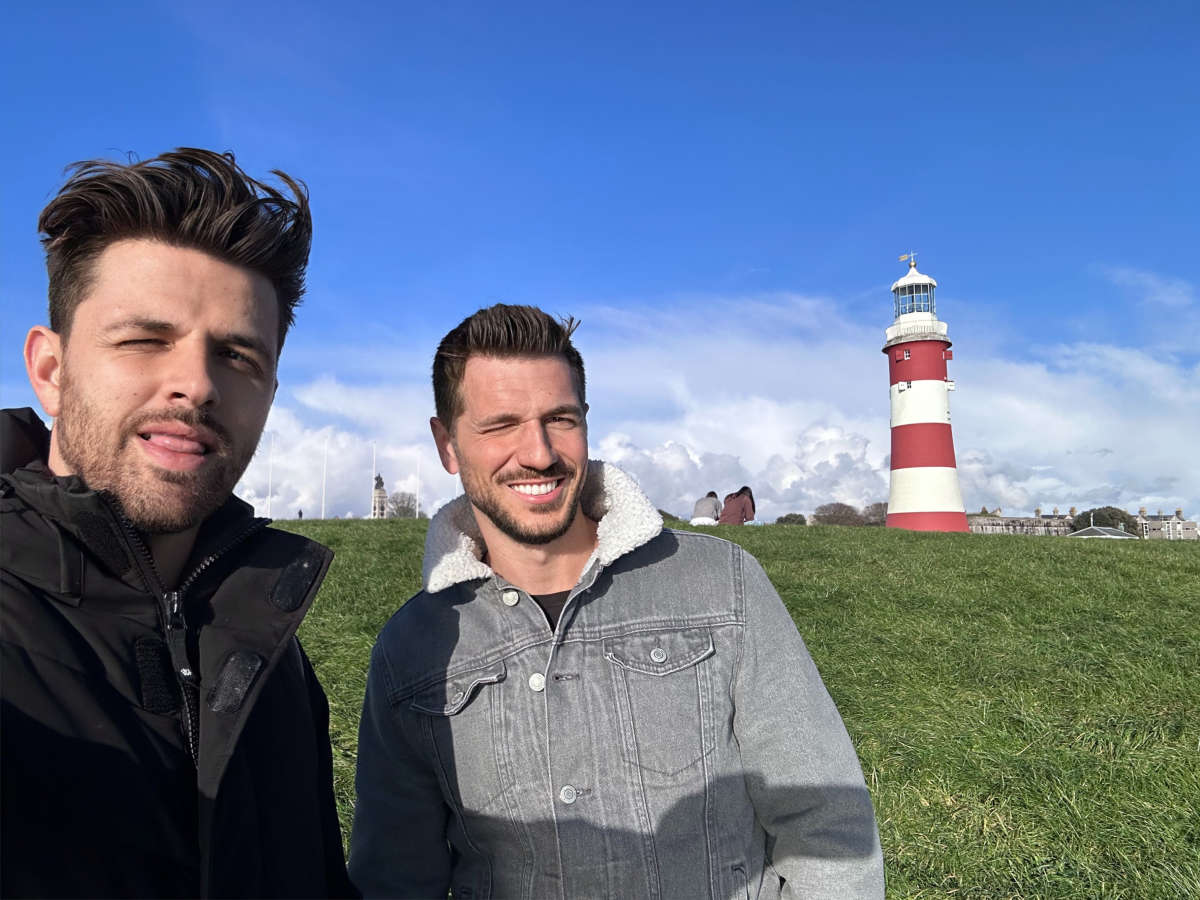 Two cheerful men taking a selfie with the iconic red and white Smeaton's Tower lighthouse in the background at Plymouth Hoe, embodying a joyful day out for friends at this historic maritime landmark.