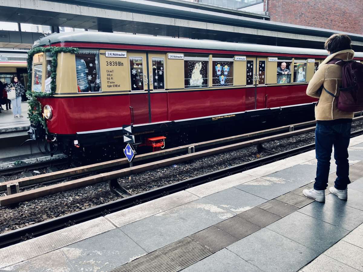 A person waiting at a Berlin S-Bahn station, observing a vintage red train carriage adorned with Christmas decorations, evoking the festive spirit of the city during the holiday season.