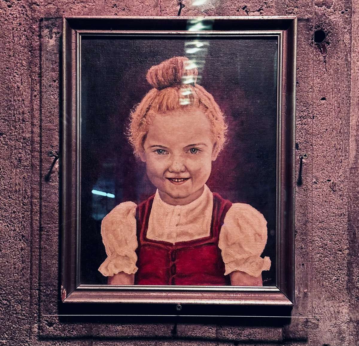 A framed painting displayed at Möbel Olfe in Berlin, depicting a young girl with a beaming smile and red hair styled in a bun, wearing a traditional red dress, adding a touch of vintage charm to the bar's eclectic decor.