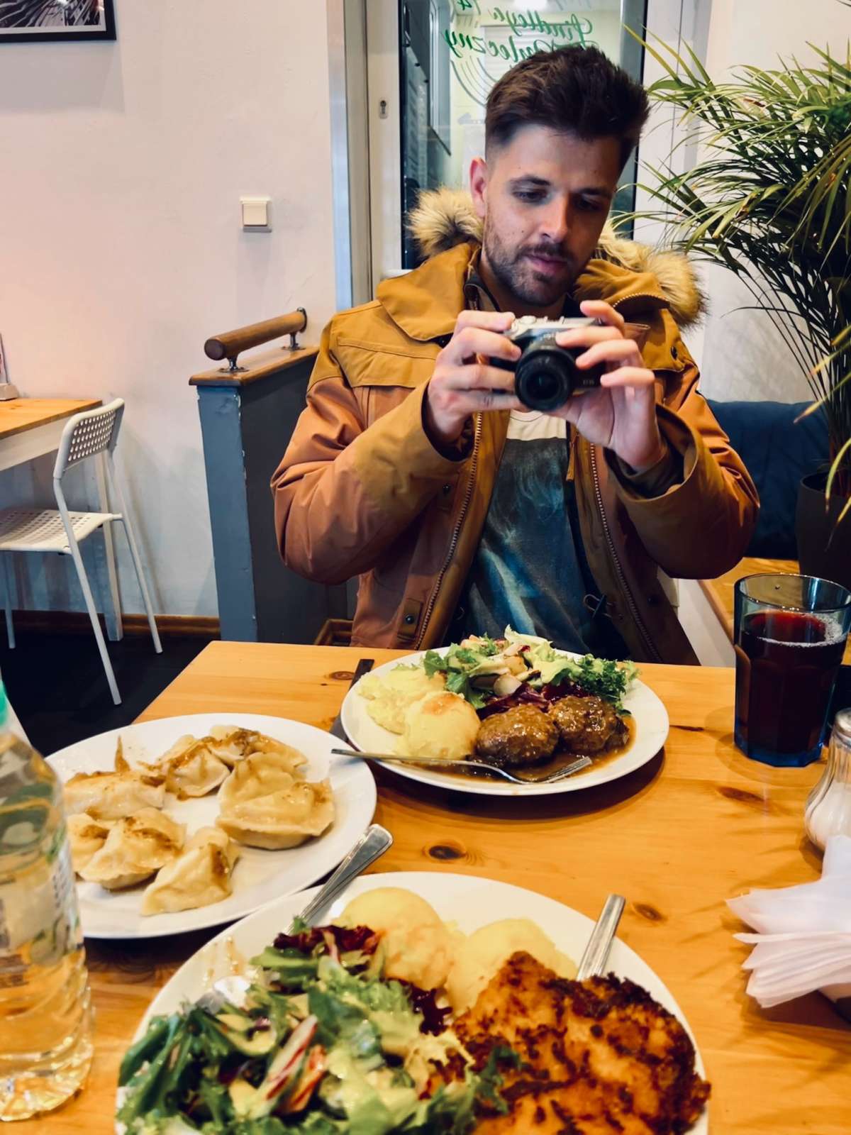 A young man with styled hair and a mustard jacket examines his camera in a cozy setting of the best milk bar in Warsaw, with plates of traditional Polish dishes like pierogi and meat with potatoes in the foreground.