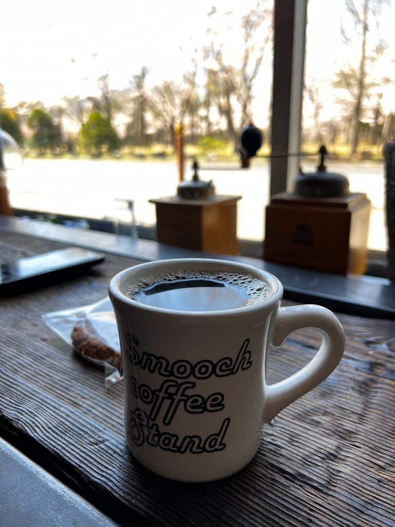 A mug of black coffee on a wooden table with 'Smooch Coffee Stand' written on it, set against a blurred park view in Sapporo, Hokkaido itinerary.