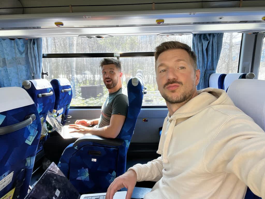 Two smiling men seated on a bus, with one working on a laptop, depicting the digital nomad lifestyle while traveling through Hokkaido itinerary.