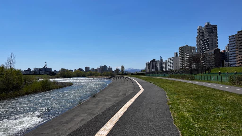 A sunny day by the Toyohira River in Sapporo with a paved walkway alongside the flowing water, with urban buildings in the distance, a pleasant route on a Hokkaido itinerary