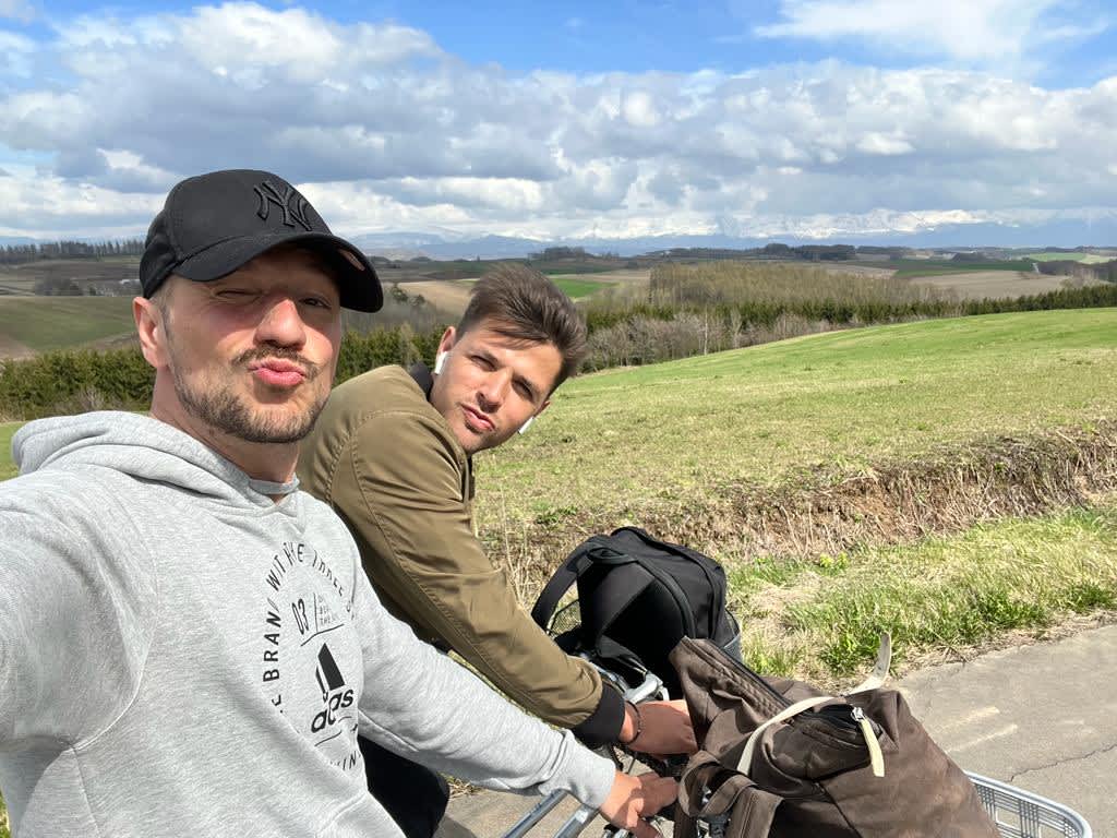 Two friends making faces at the camera with rolling green hills and distant mountains in Hokkaido, a delightful moment during a cycling tour on a Hokkaido itinerary.