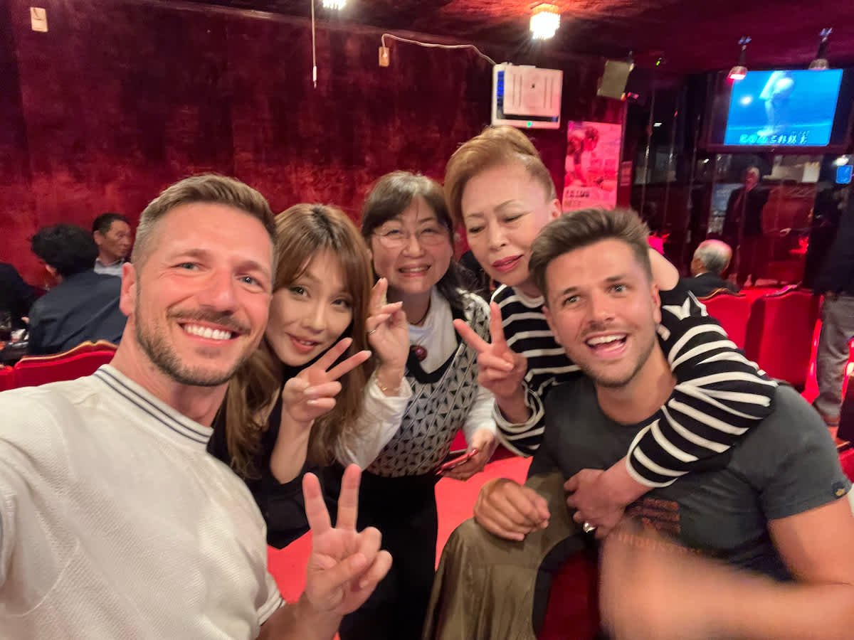 A selfie with joyful international visitors and friendly Japanese locals in a cozy bar (Snack Ringu) setting in Biei, Hokkaido, a warm cultural exchange during a Hokkaido itinerary.