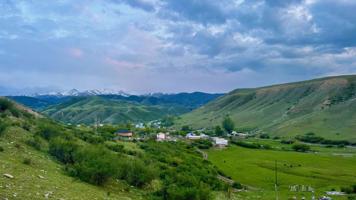 A scenic view of the lush green landscape and rolling hills in Karabulak, Kazakhstan, with a village nestled in the valley and snow-capped mountains in the background, ideal for a serene road trip.