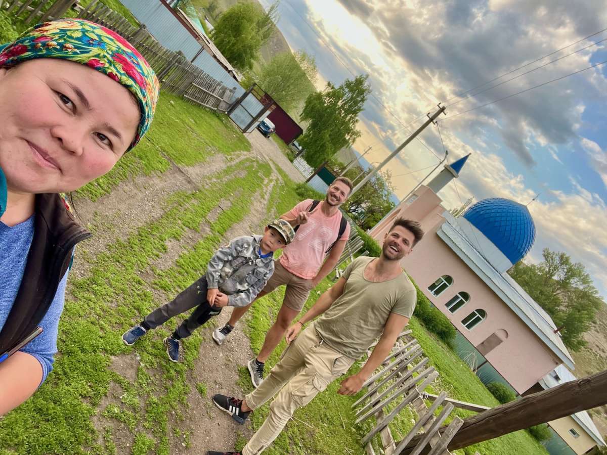 Local woman in a vibrant headscarf taking a selfie with three travelers in a rural area of Karabulak, Kazakhstan, with a traditional mosque in the background, reflecting the cultural experiences of a Kazakh road trip.