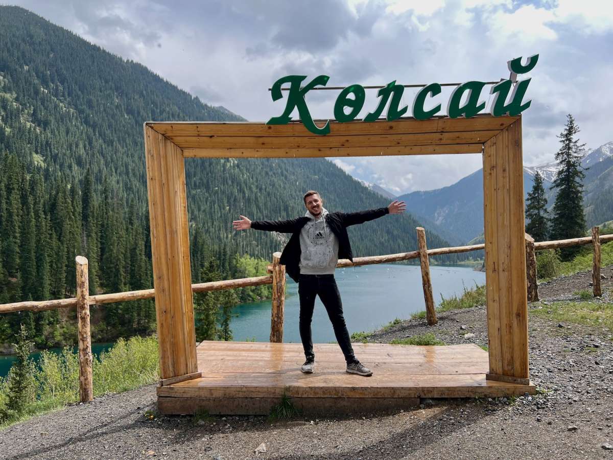 Man posing with outstretched arms in a large wooden frame with 'Колсай' written above, at Kolsai Lake, capturing the excitement of exploring the natural beauty of Kazakhstan.