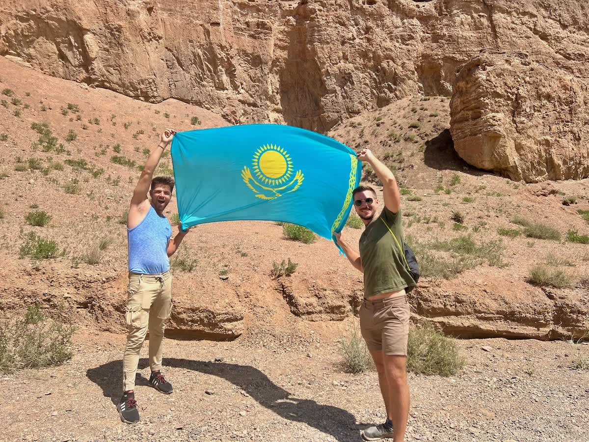 Two travelers joyfully holding the blue and yellow flag of Kazakhstan in the rugged terrain of Charyn Canyon, symbolizing national pride during a road trip adventure.