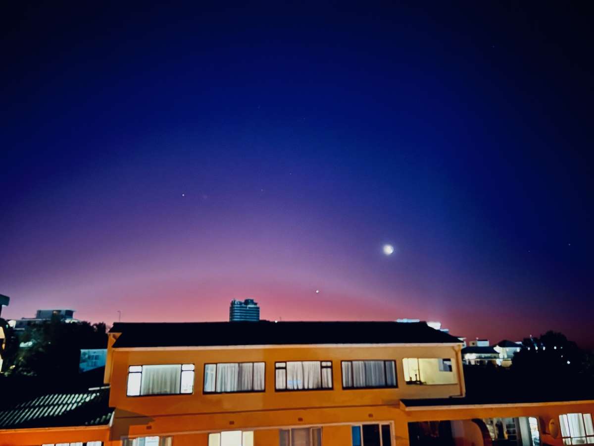 Twilight view over a residential area in gay Cape Town with gradient skies transitioning from deep blue to vivid pink, featuring a bright moon and silhouetted buildings.