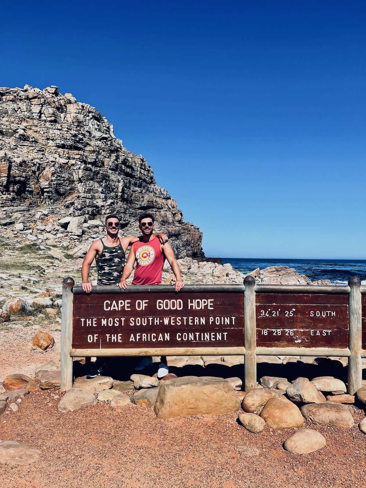 Two men smiling at the Cape of Good Hope sign, marking the most south-western point of the African continent, a popular landmark in gay Cape Town.