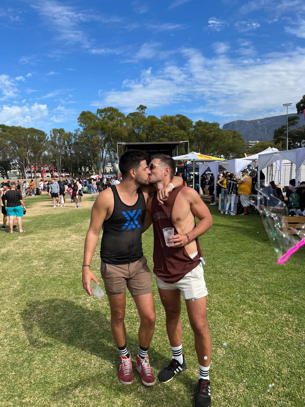 Two men sharing a kiss at Cape Town Pride, with festival-goers and flags in the background, celebrating love and diversity in the heart of South Africa.