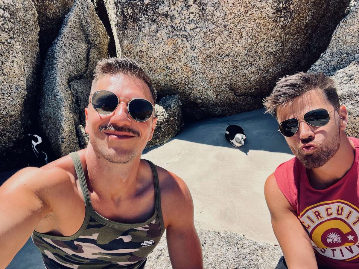 A playful selfie of two men with African penguins in the background, capturing a moment of joy and nature in gay Cape Town.